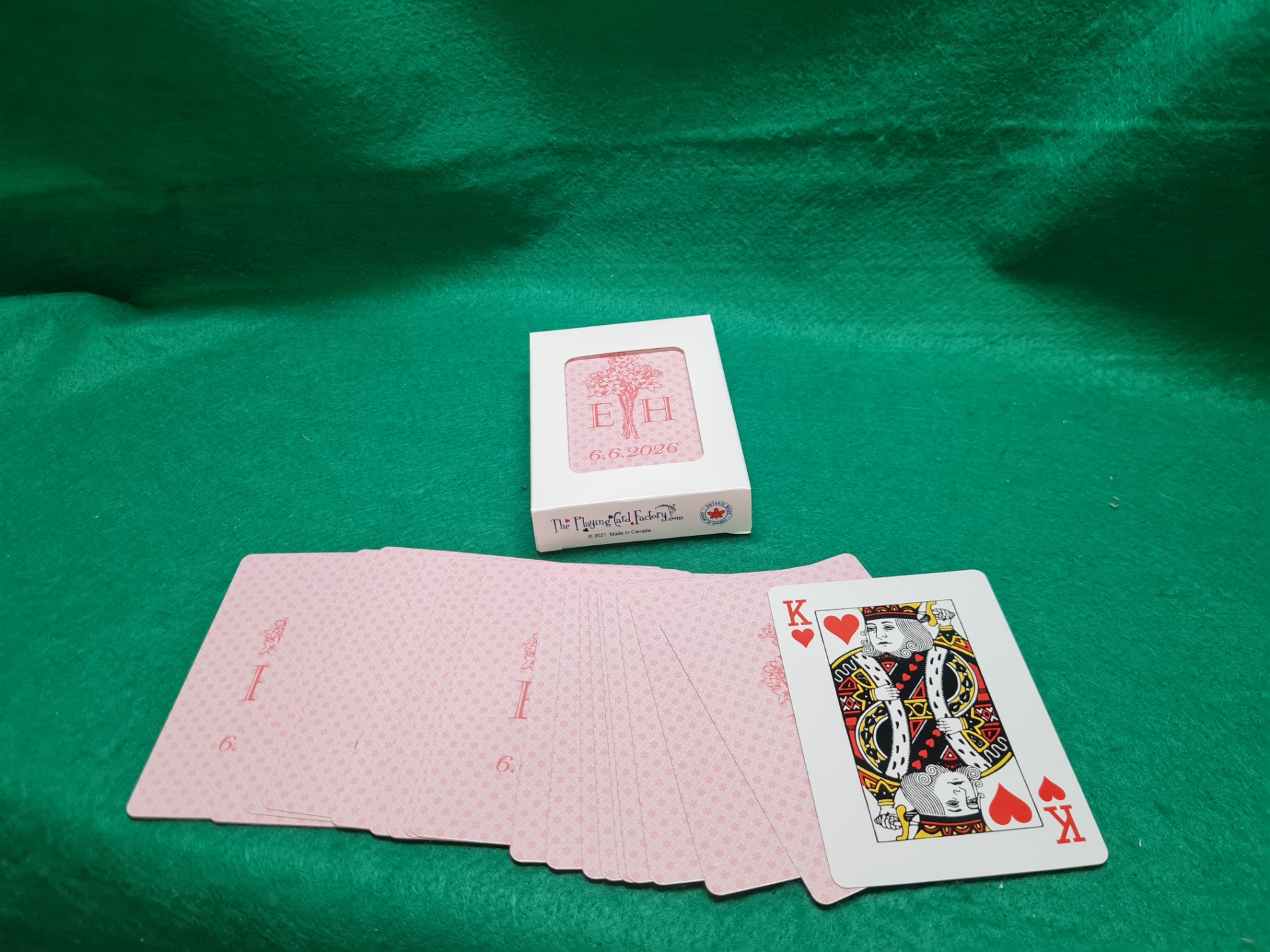 20260611_141509 Custom wedding playing cards with monogram "E H" and date "6.6.2026" on a white tuck box, displayed with the full deck fanned out showing a blush pink custom card back design, on a green felt surface. Printed by The Playing Card Factory.