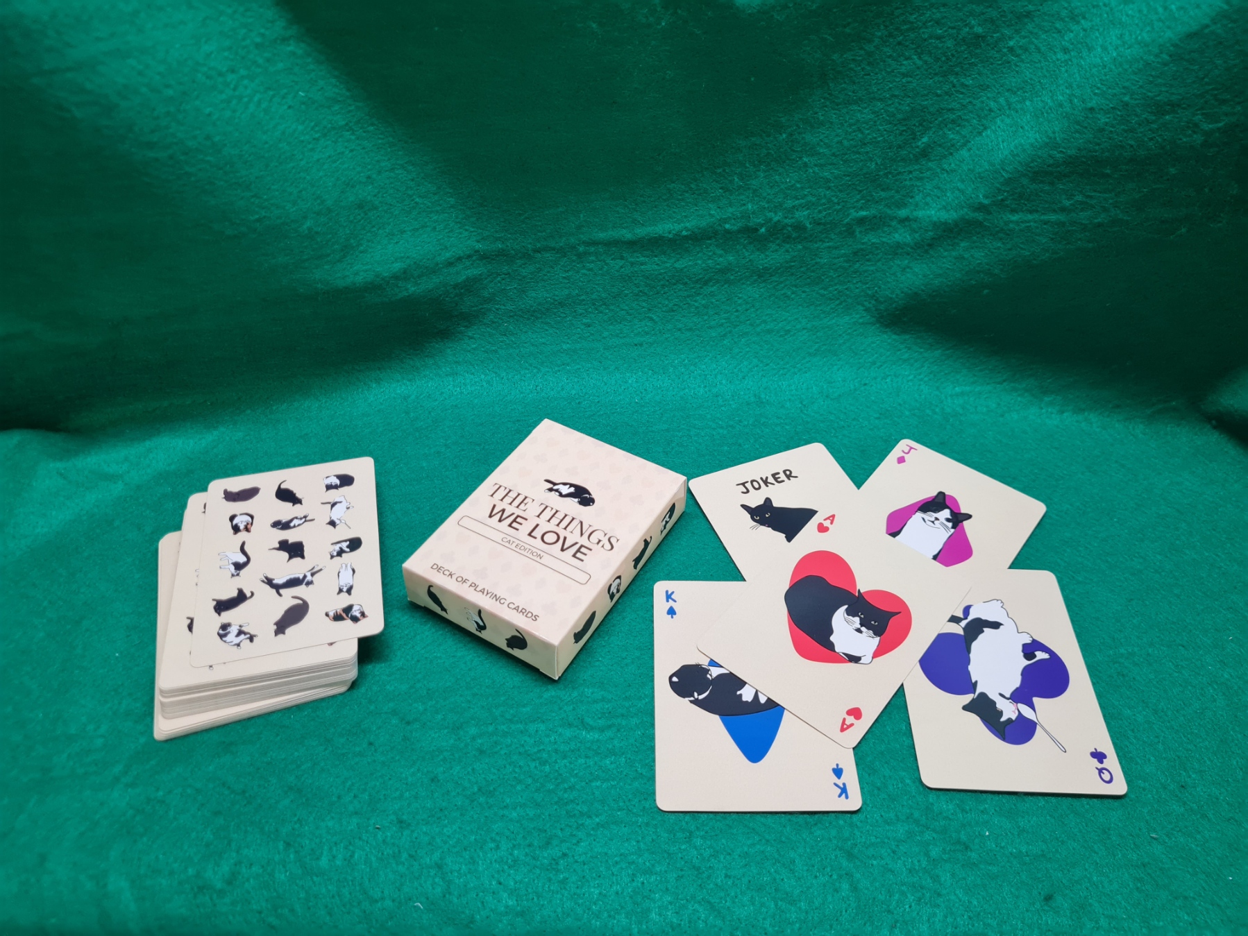 20260611_141715 Custom playing card deck titled "The Things We Love – Cat Edition" displayed on a green felt surface, showing the tuck box and five face-up cards featuring illustrated black-and-white cat artwork on cream-coloured card stock.