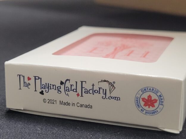 20260624_172951 Close-up of the bottom of a custom playing card tuck box showing The Playing Card Factory logo, "© 2021 Made in Canada" text, and an "Ontario Made – Fabriqué en Ontario" seal with a red maple leaf, with the blush pink card back visible through the window above.