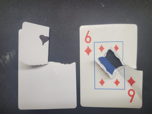 Two playing cards on a dark surface — a torn 9 of Diamonds revealing the dark black core layer found in The Playing Card Factory's premium card stock, compared to a torn blank card showing the white-through stock used by lower-quality manufacturers, demonstrating why black core is the professional standard.