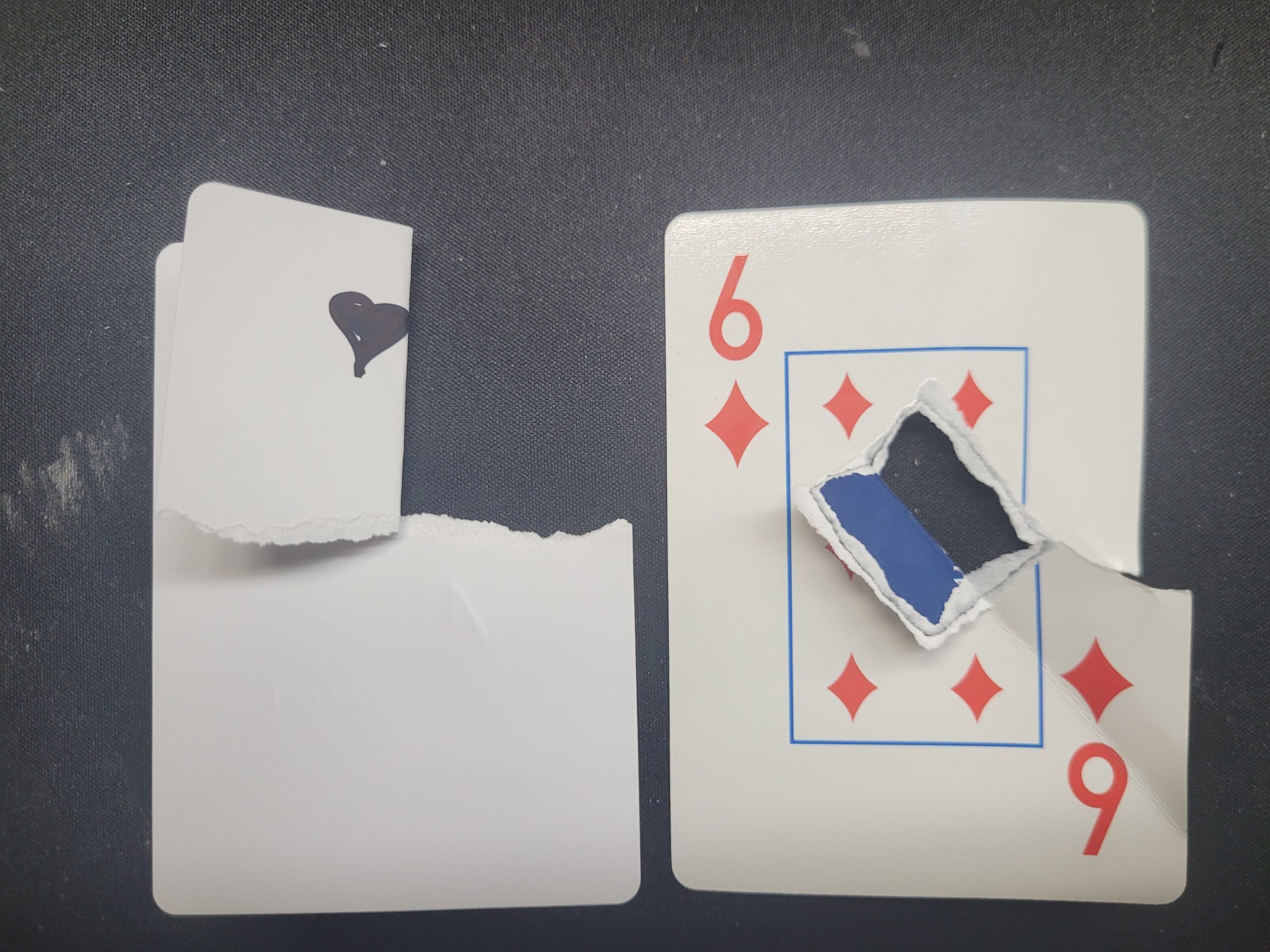 Two playing cards on a dark surface — a torn 9 of Diamonds revealing the dark black core layer found in The Playing Card Factory's premium card stock, compared to a torn blank card showing the white-through stock used by lower-quality manufacturers, demonstrating why black core is the professional standard.