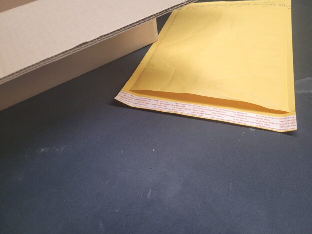 A gold kraft bubble mailer with peel-and-seal adhesive strip shown alongside a double-walled corrugated cardboard shipping box, used for packaging custom playing card orders at The Playing Card Factory.
